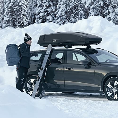 Person loading skis onto a car with a roof box in snowy landscape.